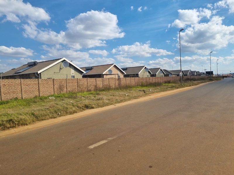 A street-level view of a residential development featuring a row of houses with tiled roofs, solar geysers, and a secure perimeter brick wall.
