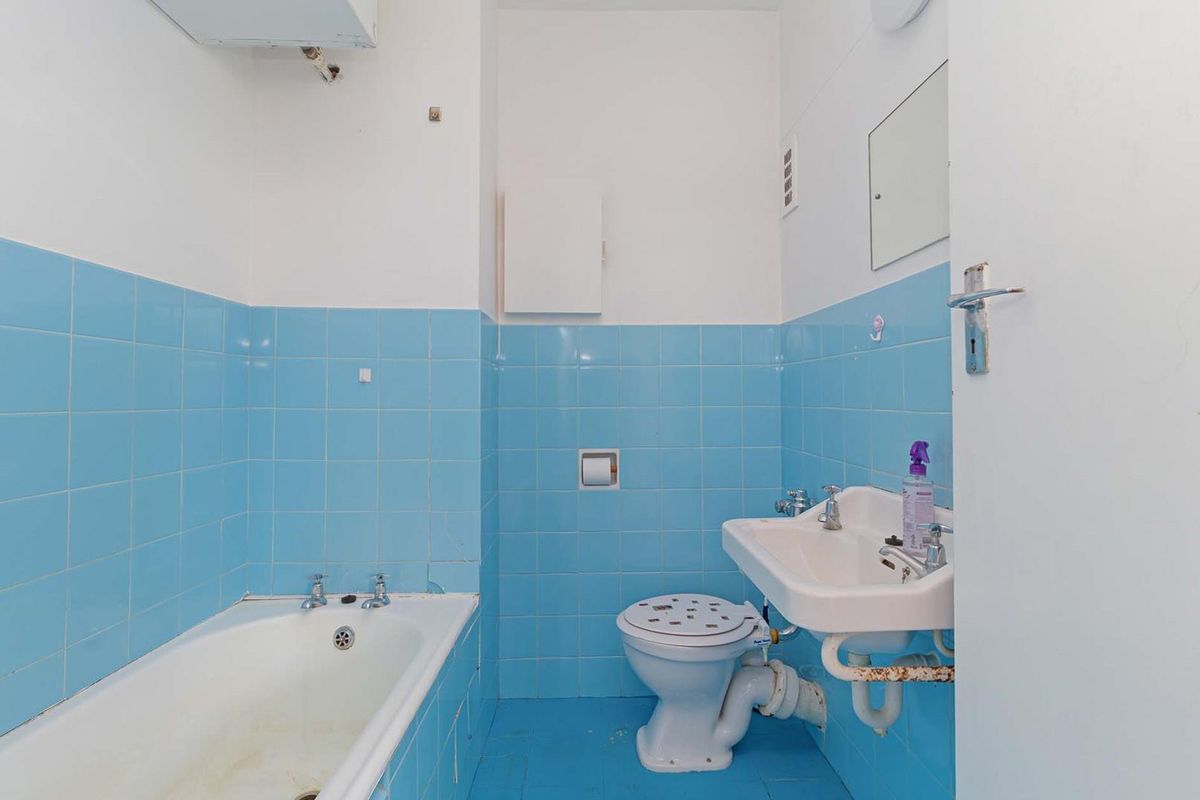 A dated bathroom featuring blue wall and floor tiles, a standard bathtub, pedestal basin, and toilet, requiring cosmetic updates and plumbing maintenance.