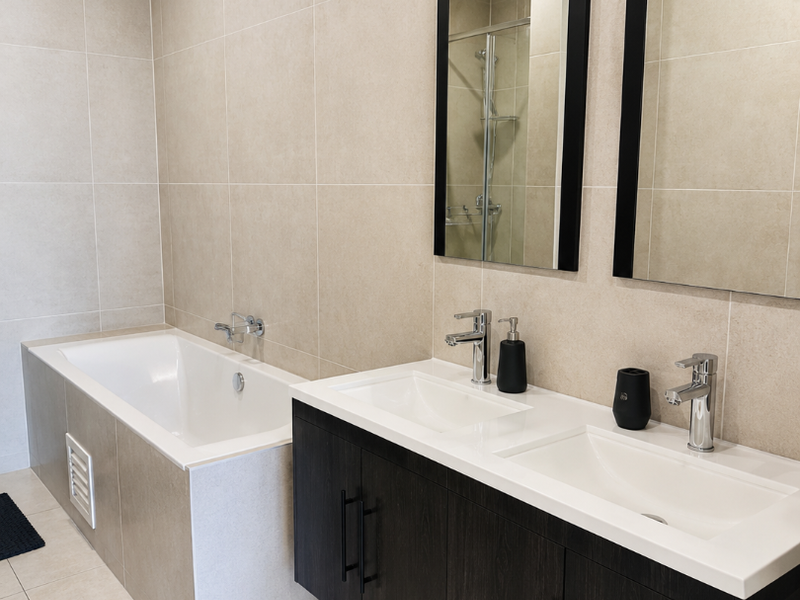 Modern bathroom featuring a double vanity with dark cabinetry, large wall-mounted mirrors, and a tiled built-in bathtub with neutral wall and floor tiles.
