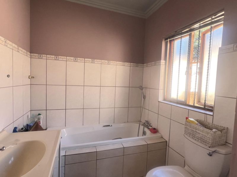 A functional bathroom featuring a built-in bathtub, pedestal basin, and toilet, finished with classic wall tiling and a window with security bars.