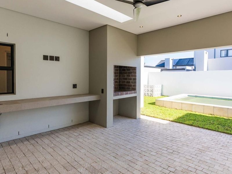 Covered outdoor patio featuring a built-in braai, stone prep counter, and ceiling fan, overlooking a private garden and swimming pool area.