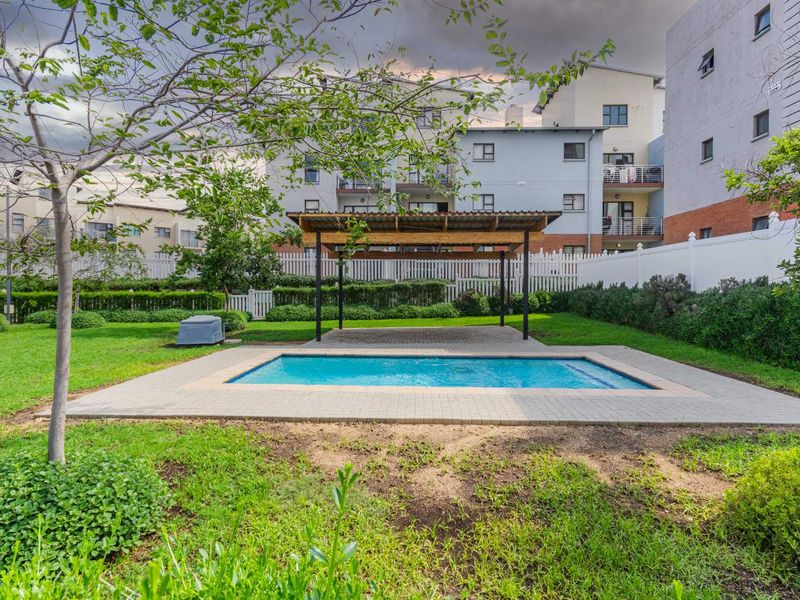 Communal outdoor area featuring a swimming pool, a covered pergola, and landscaped garden surrounds within an apartment complex.
