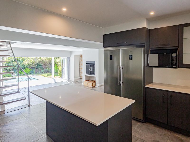 Modern open-plan kitchen featuring dark cabinetry, stone countertops, and a central island, seamlessly flowing into a living area with a fireplace and staircase.
