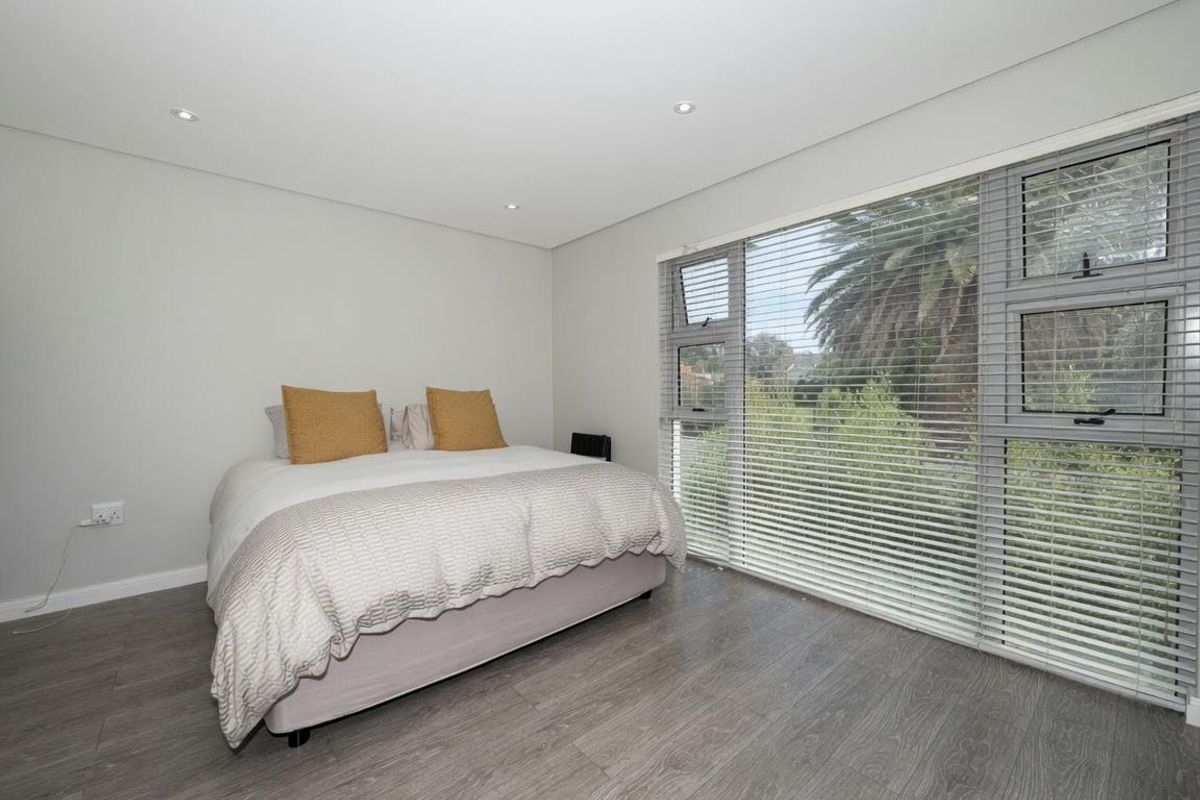 A bright and modern bedroom featuring wood-look laminate flooring, recessed ceiling lighting, and large windows with fitted horizontal blinds.