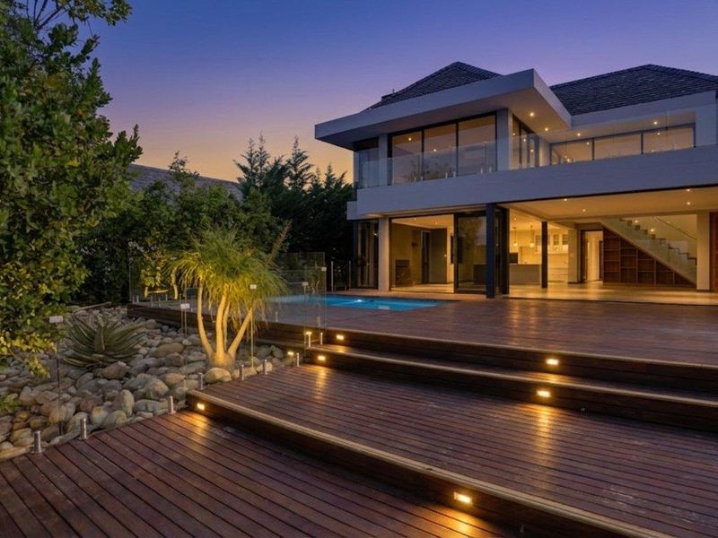 Modern double-story residence featuring a spacious timber deck, integrated swimming pool, glass balustrades, and seamless indoor-outdoor flow through large sliding glass doors.