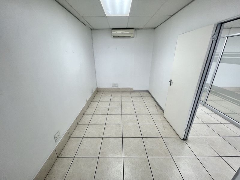 A vacant, functional office space featuring tiled flooring, a drop ceiling with recessed lighting, and a wall-mounted air conditioning unit.