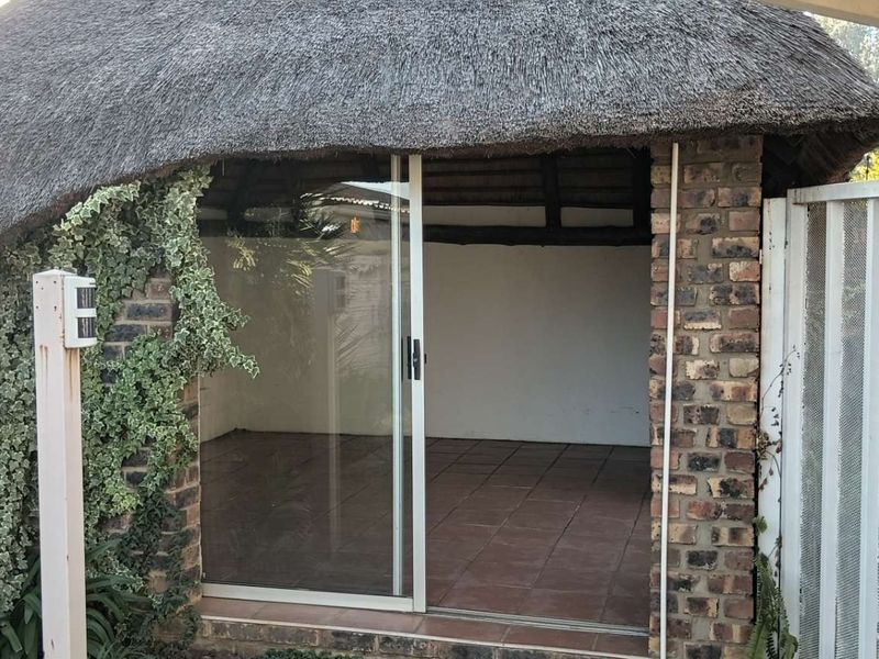 A covered outdoor patio area featuring a traditional thatch roof, exposed brick walls, and tiled flooring, accessible via a sliding glass door.