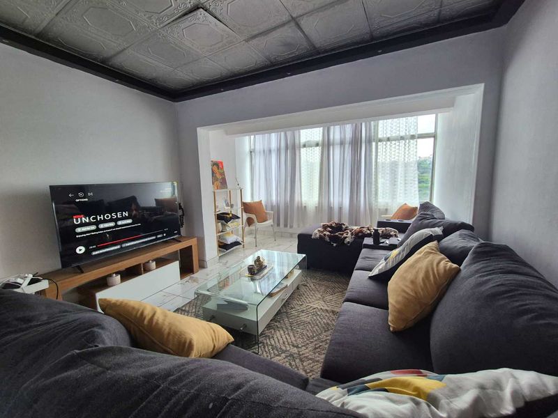 A living room featuring a distinctive pressed metal ceiling, tiled flooring, and a large window providing natural light.