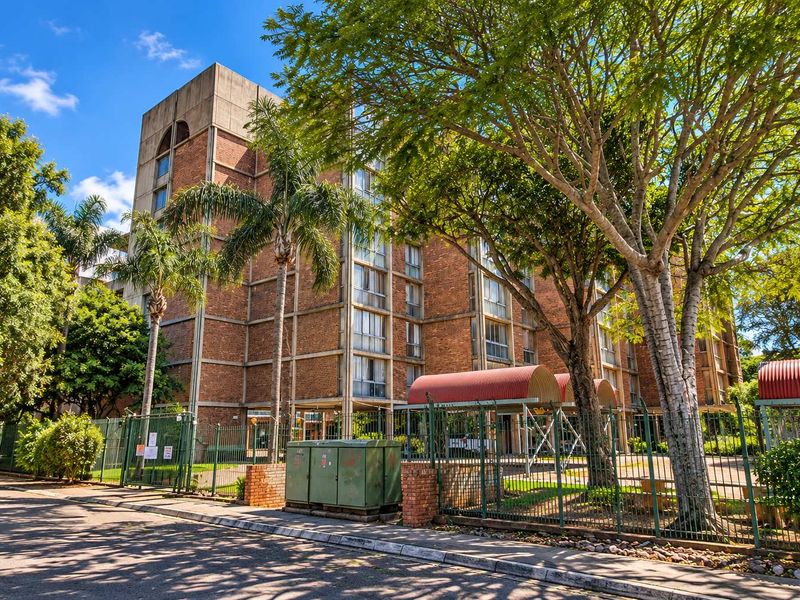 Street-level view of a multi-story brick apartment building featuring a secure fenced perimeter and arched entrance canopy.