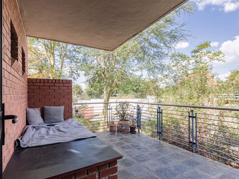 A covered balcony featuring brick wall accents, tiled flooring, and a metal safety railing overlooking mature trees.