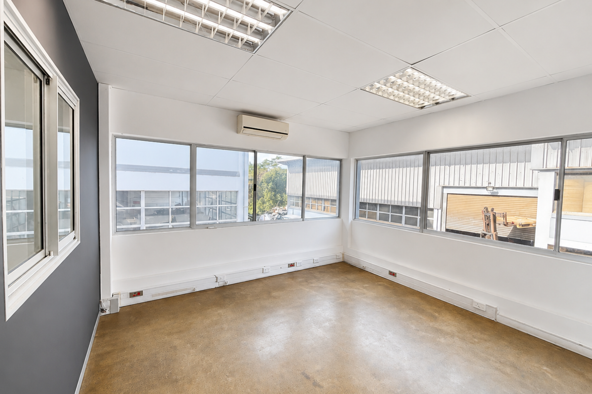 A bright, empty industrial office space featuring large windows, suspended ceiling lights, a wall-mounted air conditioning unit, and durable flooring.
