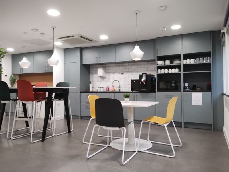 A modern office kitchen area featuring sleek grey cabinetry, subway tile backsplash, recessed lighting, and integrated sink facilities.
