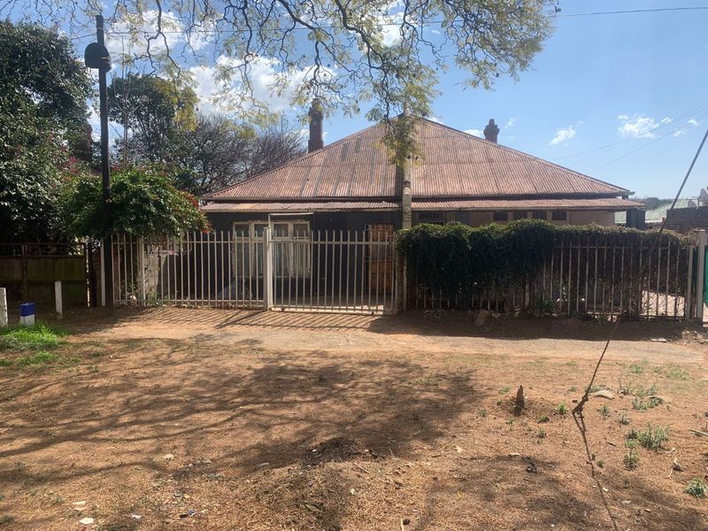 Front elevation of a single-story house featuring a corrugated metal roof, chimney stacks, and secure metal perimeter fencing.