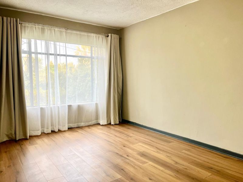 A bright, empty bedroom featuring laminate wood flooring, neutral walls, and a large window with curtain fixtures.