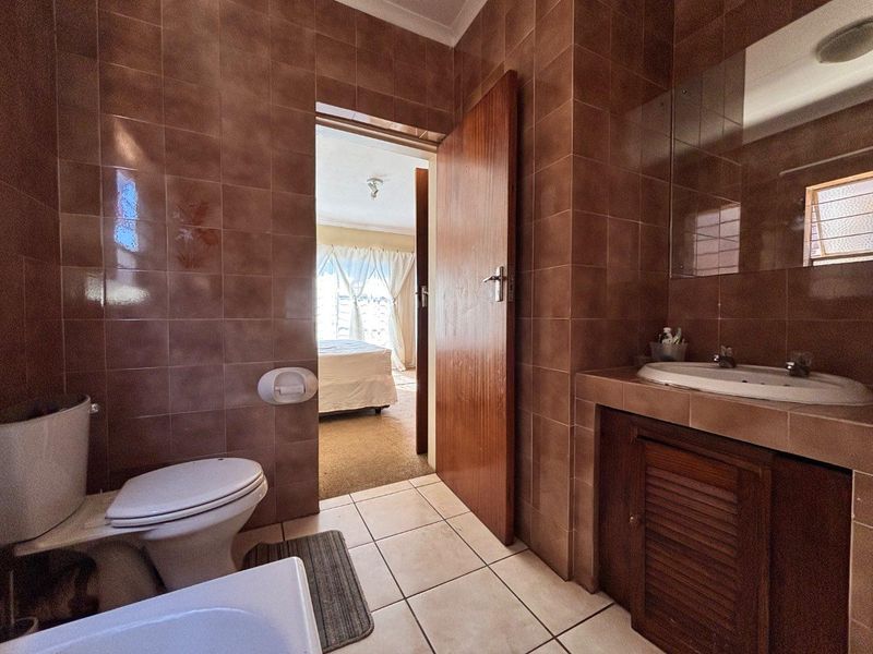 A functional bathroom featuring brown ceramic wall tiles, a built-in vanity with a washbasin, and tiled flooring, with direct access to an adjacent bedroom.