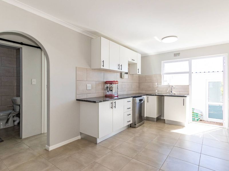 A clean, modern kitchen featuring white cabinetry, stone countertops, tiled backsplash, and direct access to an outdoor area, with an adjacent bathroom.