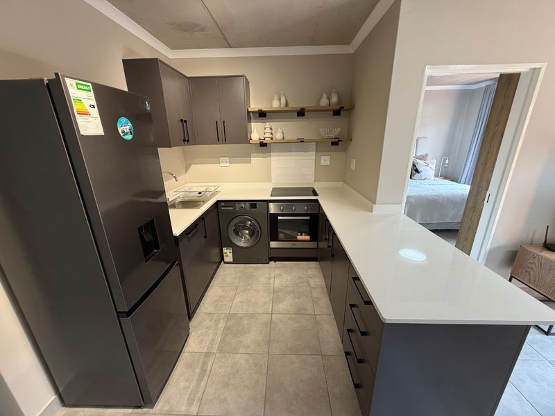 Modern kitchen featuring dark cabinetry, quartz countertops, built-in oven, electric hob, and integrated washing machine, with a view into the adjacent bedroom.