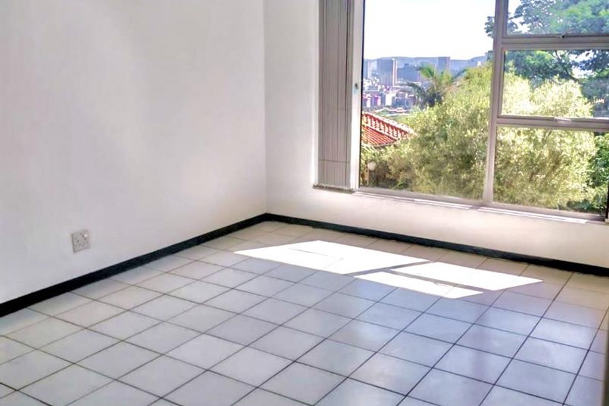 A bright, empty bedroom featuring white tiled flooring, a large window with natural light, and a view of the surrounding area.