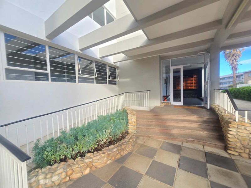 A well-maintained apartment building entrance featuring a stone-clad planter, tiled walkway, and a covered pergola structure leading to the main glass door.
