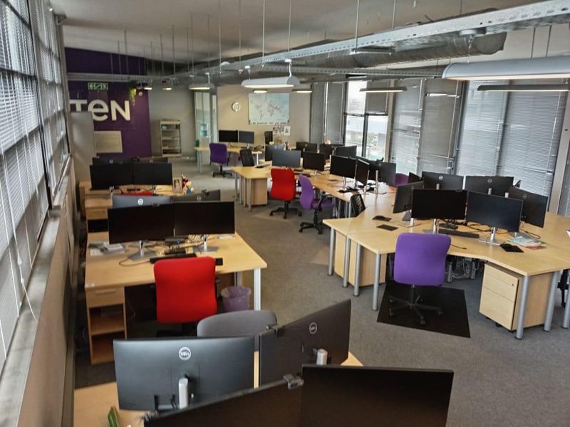 Open-plan office space featuring exposed industrial ductwork, suspended lighting, large windows with venetian blinds, and carpeted flooring.
