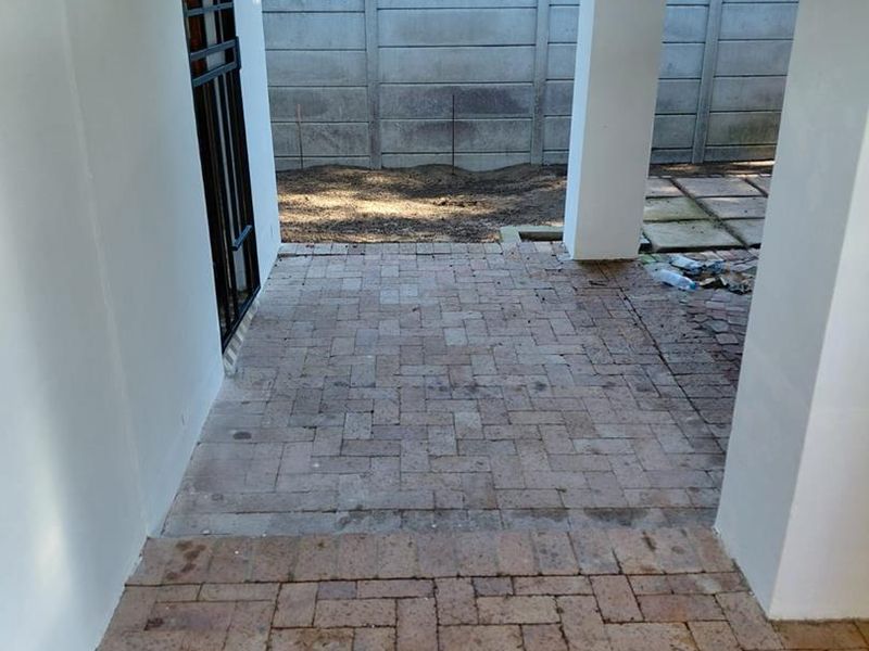 A covered outdoor patio area featuring brick paving, concrete support pillars, and a security gate, leading to a rear garden space with boundary walling.