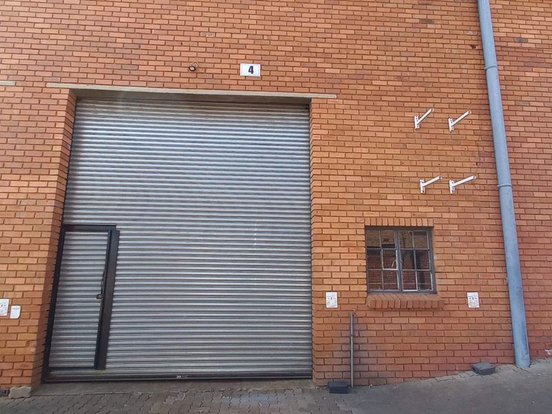 Industrial unit exterior featuring red brick facade, secure roller shutter doors, and integrated pedestrian access doors with small steel-framed windows.