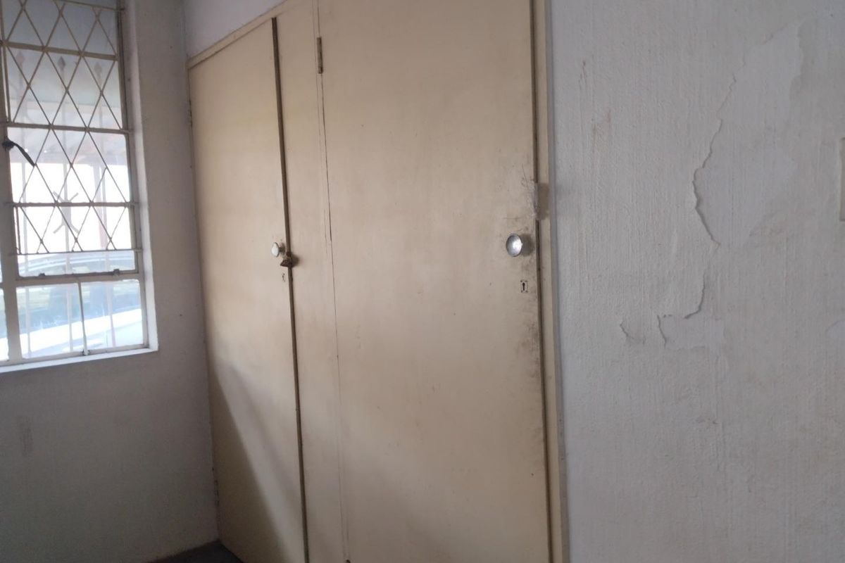 A bedroom interior featuring built-in cupboards and a window with security bars, showing signs of wear and peeling paint on the walls.