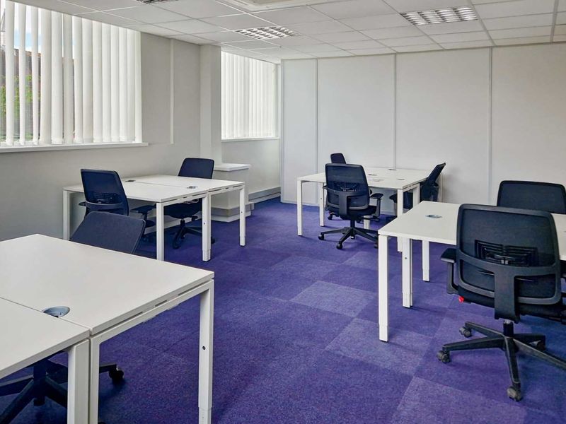 A functional office space featuring suspended ceilings, recessed lighting, vertical window blinds, and durable carpet tiles, suitable for a professional work environment.