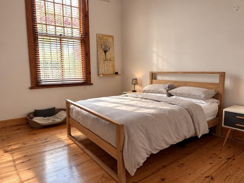 A bright and airy bedroom featuring polished timber flooring and a large sash window with timber frames, providing ample natural light.