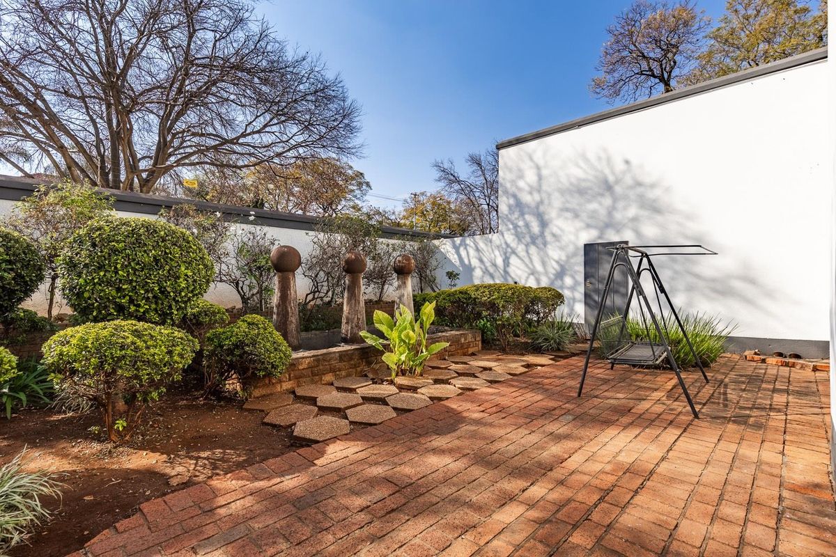 A well-maintained rear courtyard featuring brick paving, established garden beds, and decorative stone pillars against a clean white boundary wall.