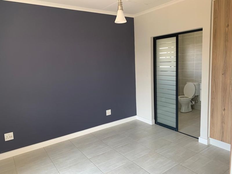 A clean, modern bedroom featuring tiled floors, a dark accent wall, built-in cupboards, and an en-suite bathroom with a sliding door.