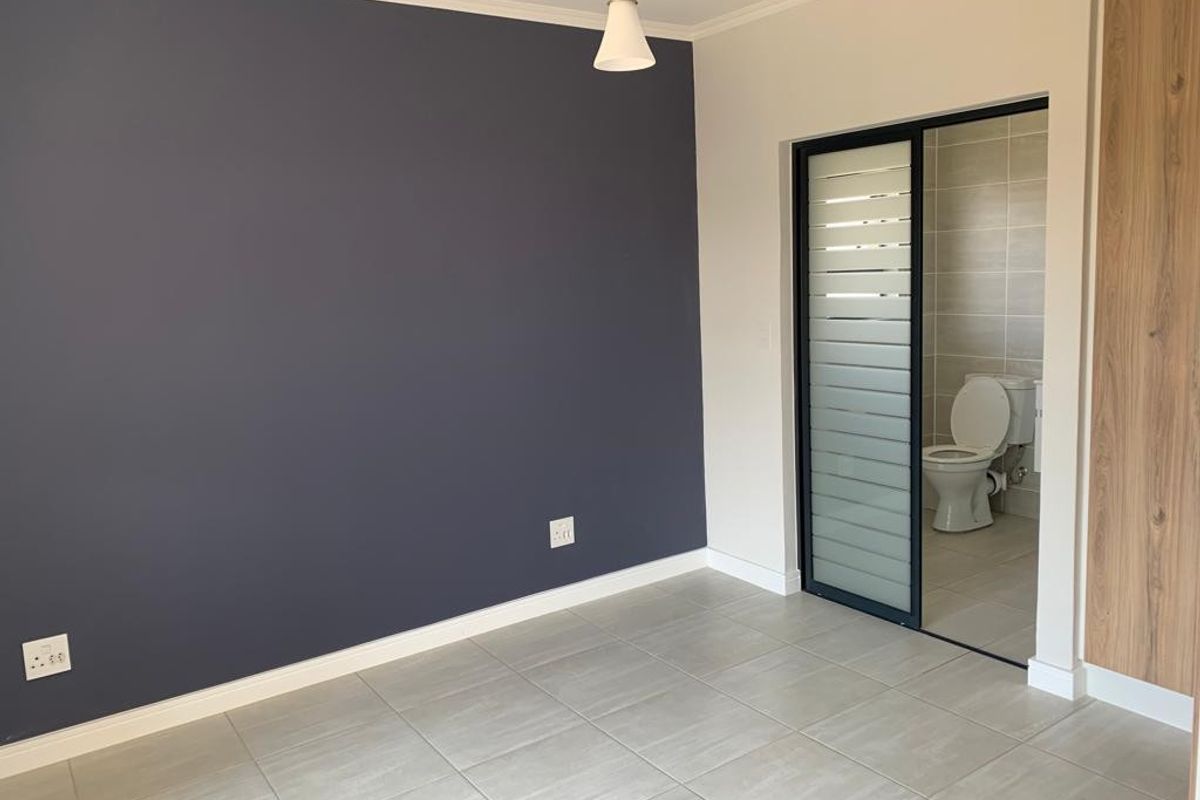 A clean, modern bedroom featuring tiled floors, a dark accent wall, built-in cupboards, and an en-suite bathroom with a sliding door.