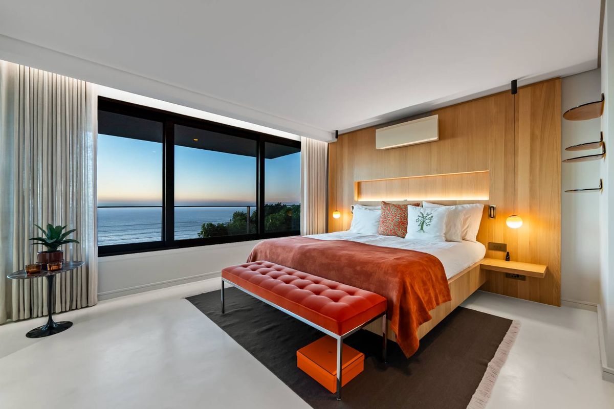 A modern, high-end bedroom featuring warm timber wall paneling, integrated lighting, and expansive floor-to-ceiling windows offering stunning ocean views.