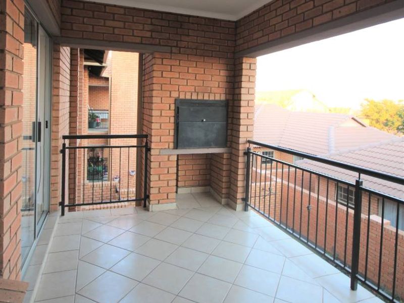 A spacious covered balcony featuring exposed brick walls, tiled flooring, a built-in braai, and metal balustrades, offering a functional outdoor entertainment area.