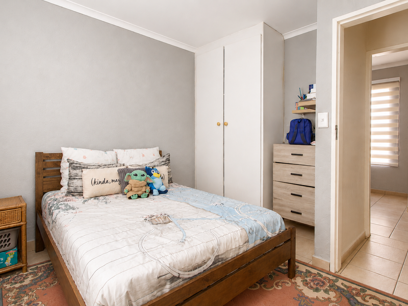 A functional bedroom featuring a built-in wardrobe, neutral wall finishes, and tiled flooring, with an adjacent hallway view.