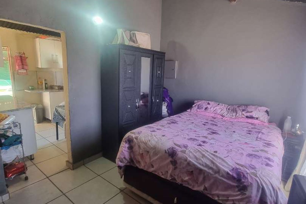 A bedroom featuring tiled flooring and an exposed corrugated metal roof, with an adjacent view into a kitchen area.