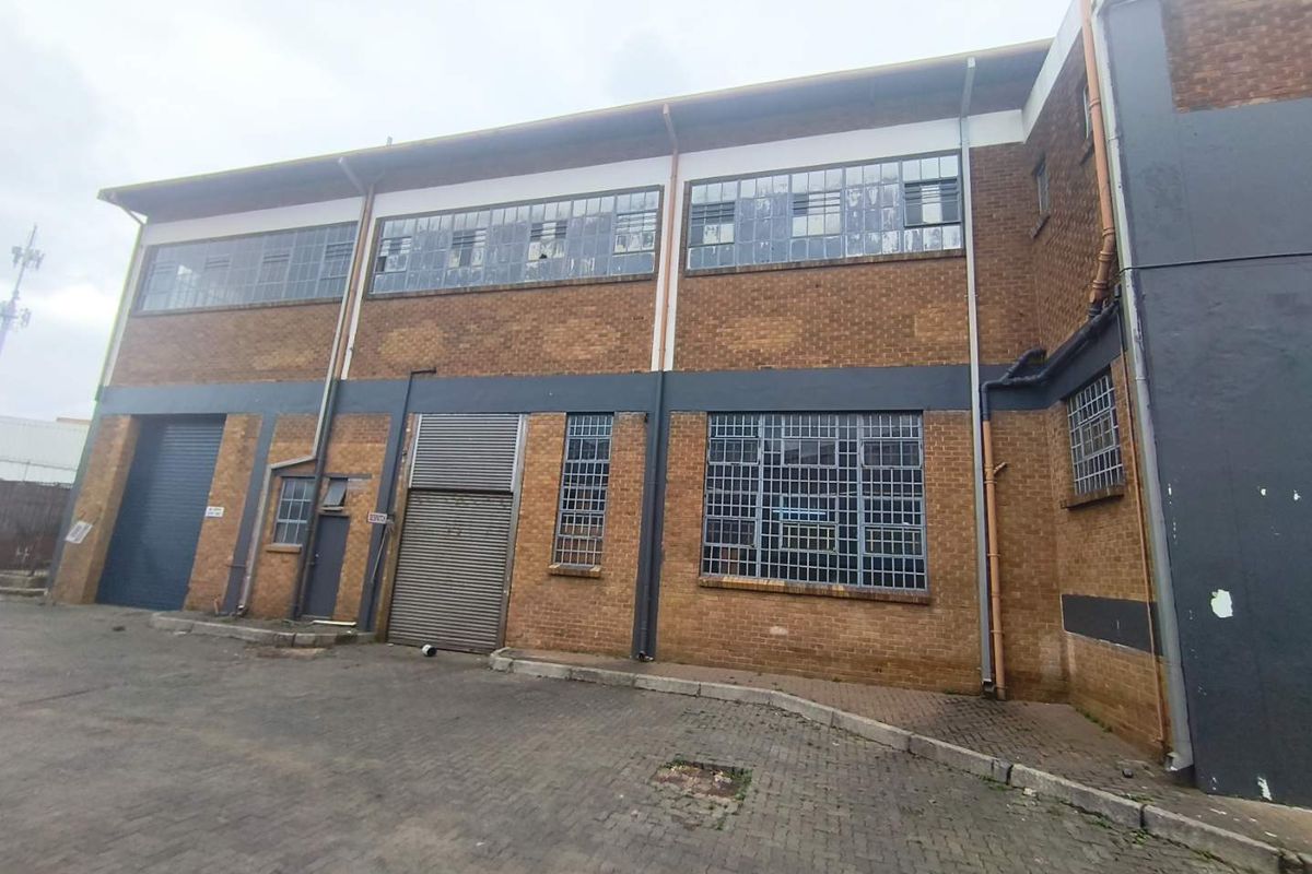 Industrial building exterior featuring brick facade, steel-framed windows, and multiple roller shutter access points with paved driveway area.