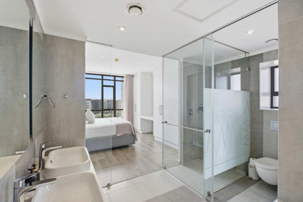 Modern en-suite bathroom featuring a glass-enclosed shower, dual vanity basins, and contemporary tiling, seamlessly integrated with the adjacent bedroom area.