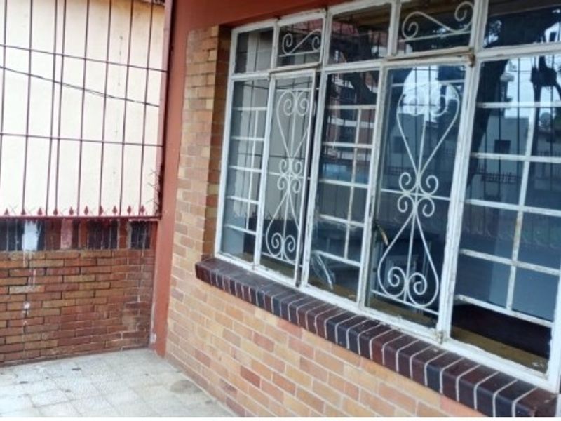 A covered patio area featuring exposed brick walls, tiled flooring, and security-barred windows, offering a functional outdoor space in need of maintenance.