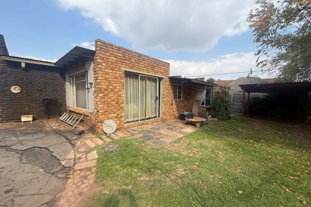 Rear exterior view of the house featuring exposed brick facade, sliding glass doors, paved patio area, and a garden space with a carport structure.