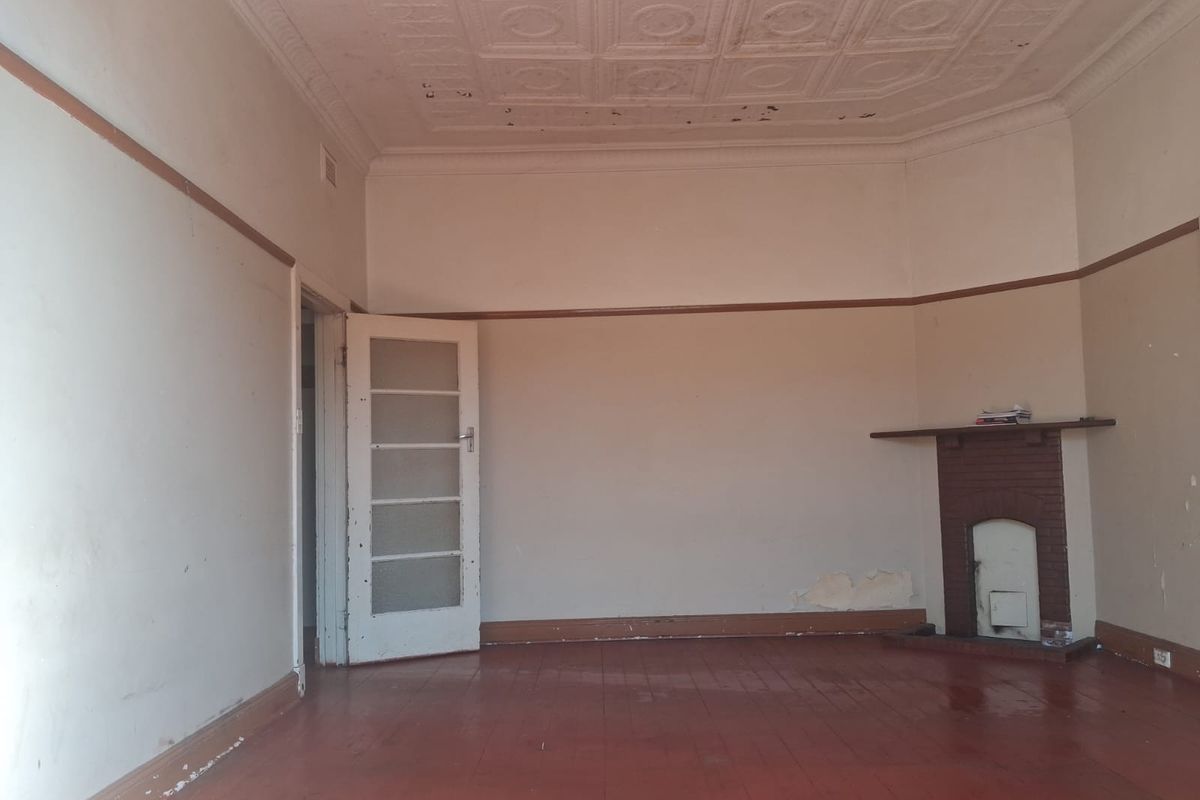 An empty living room featuring a classic pressed metal ceiling, original timber flooring, and a brick fireplace, requiring cosmetic renovation to restore its character.