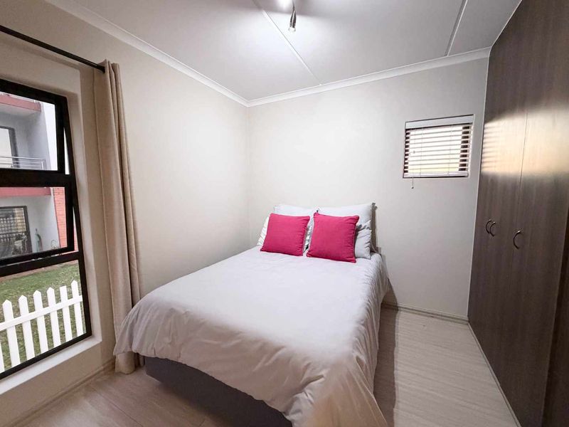 A neat bedroom featuring built-in cupboards, light laminate flooring, and large aluminium windows with venetian blinds, offering a clean and functional living space.