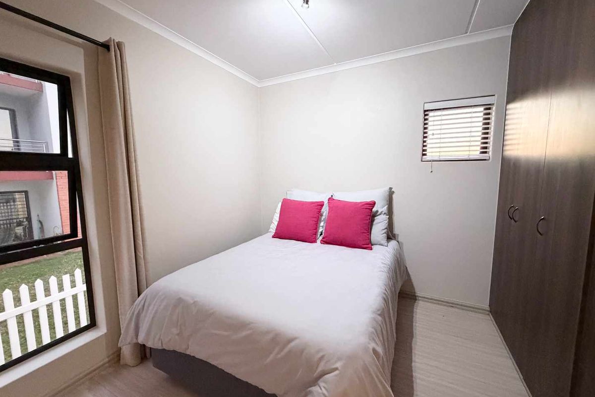 A neat bedroom featuring built-in cupboards, light laminate flooring, and large aluminium windows with venetian blinds, offering a clean and functional living space.