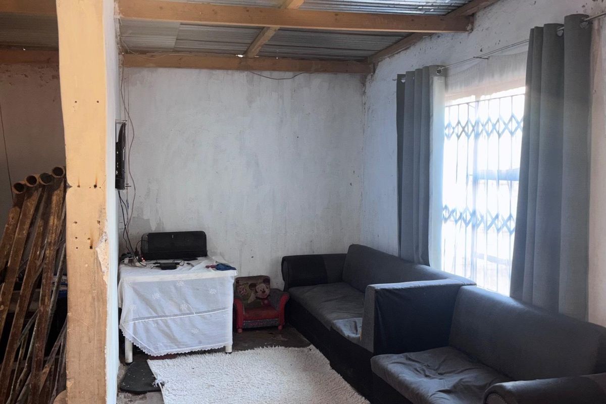 A living room area featuring exposed timber ceiling beams, corrugated metal roofing, and security gate window protection, requiring cosmetic updates to improve the interior finish.