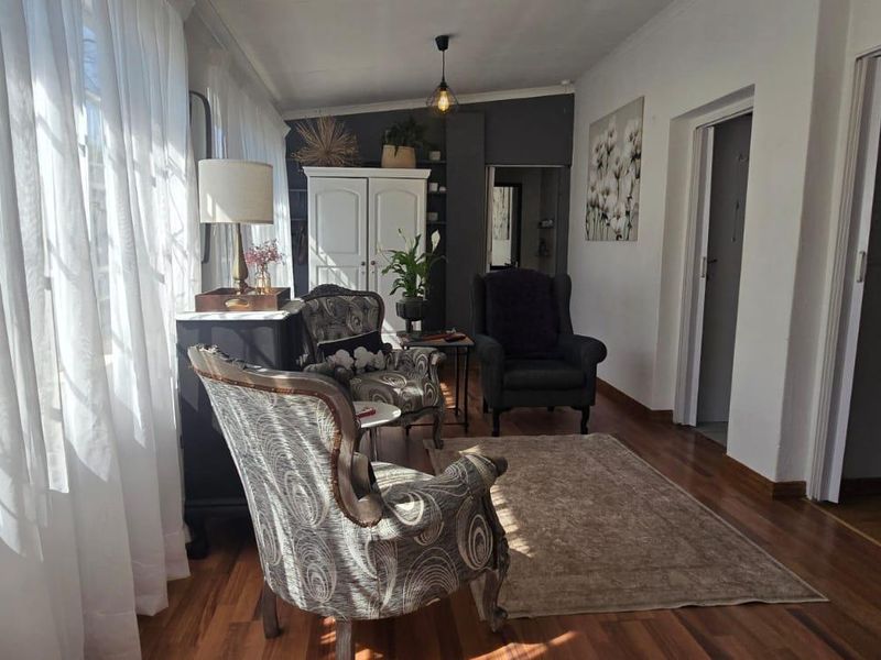 A bright living room featuring polished hardwood flooring, cove cornicing, and a built-in cupboard, illuminated by natural light through large windows.