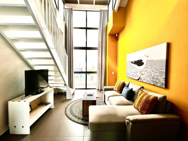A bright living room featuring double-volume ceilings, large floor-to-ceiling windows, and a modern staircase leading to an upper level.