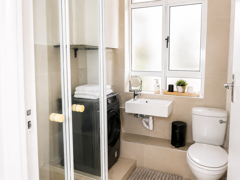 A compact, modern bathroom featuring a shower enclosure, wall-mounted basin, and toilet with neutral wall tiling and natural light from a window.