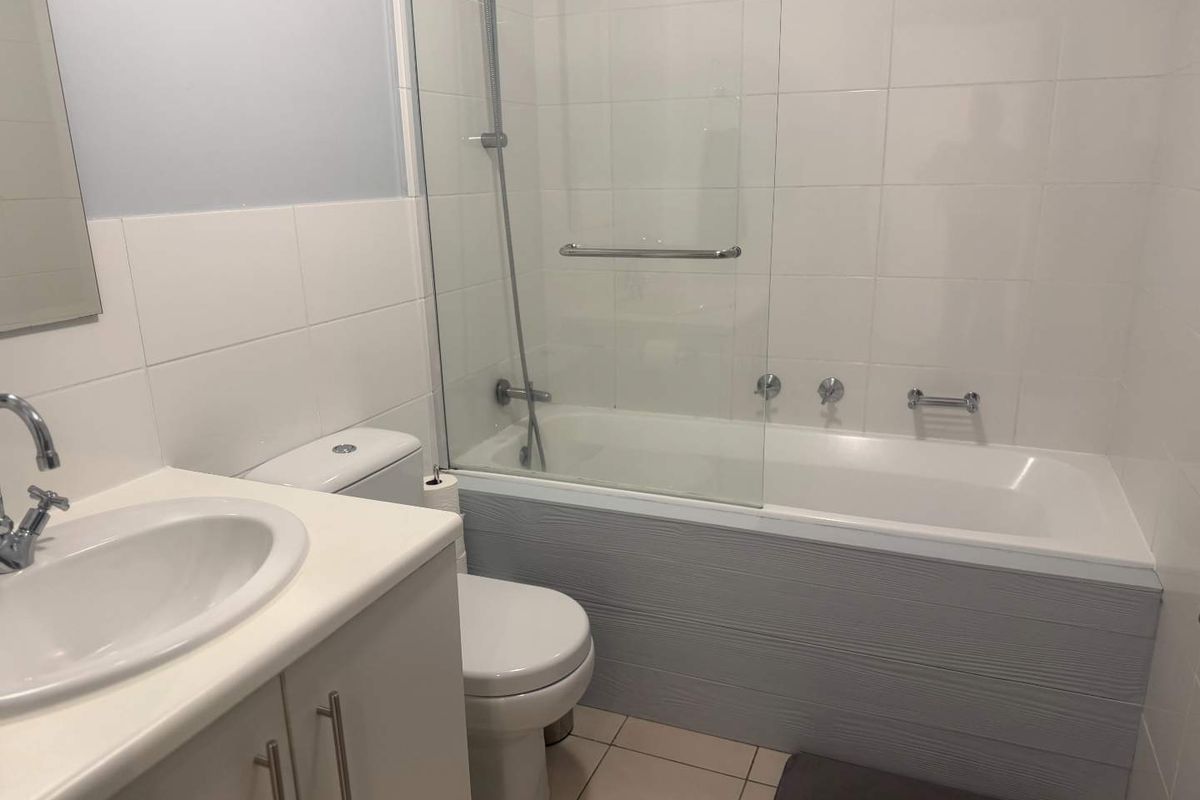 A functional bathroom featuring a white vanity, a combined shower and bathtub with a glass screen, and neutral wall tiling.