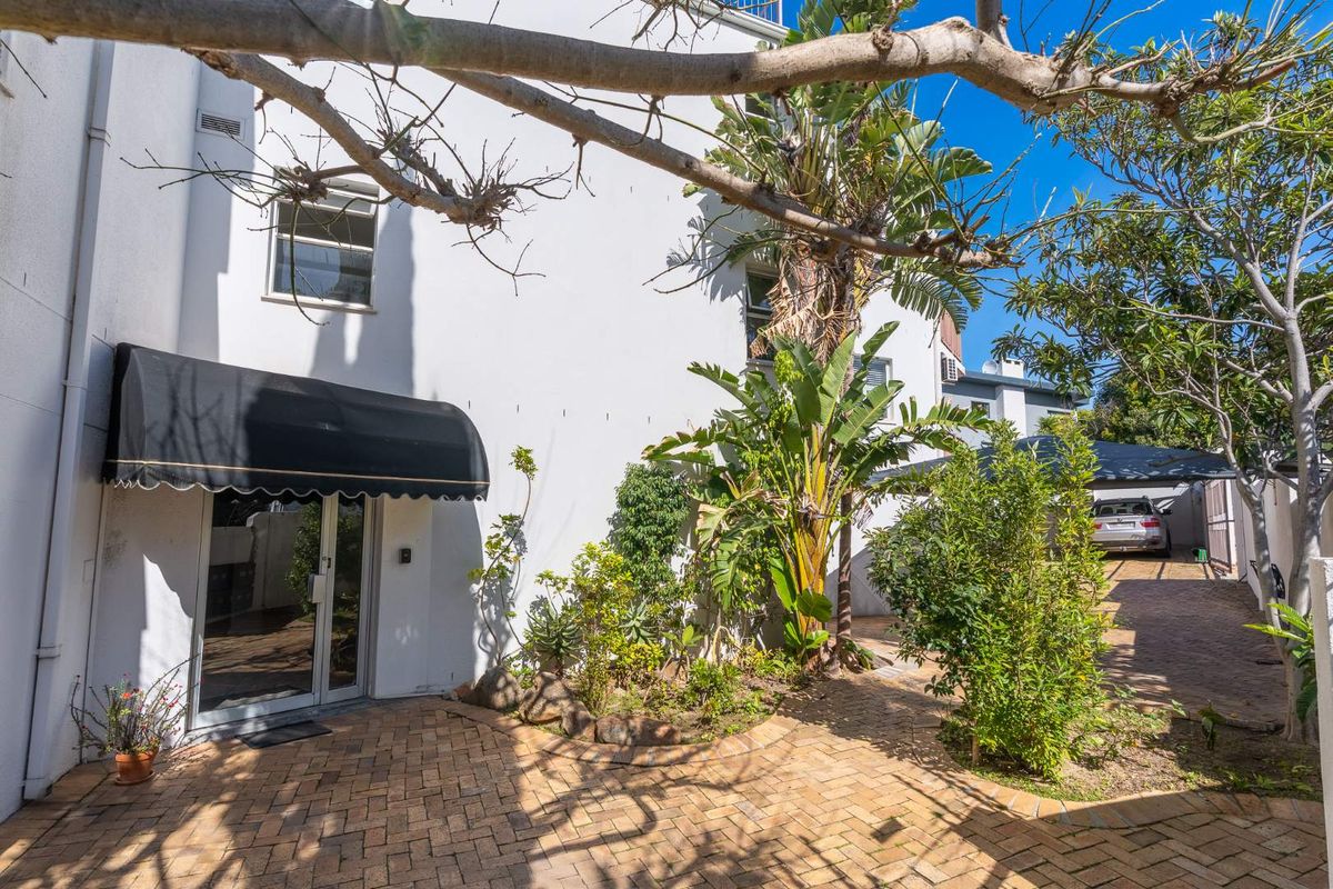 Rear exterior view of the apartment featuring a paved courtyard, established garden, and a covered entrance with a black awning.