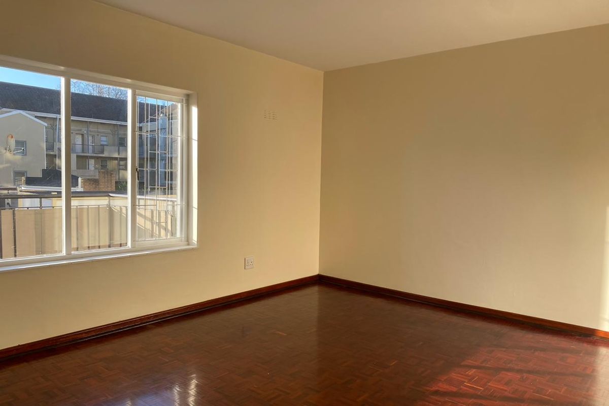 A bright, empty bedroom featuring polished parquet flooring, neutral walls, and a large sliding window with security bars.